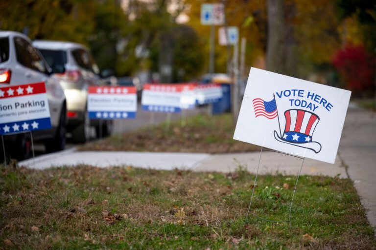 A grassy neighborhood sidewalk with cars parked and yard signs in the background on the left side and a white yard sign with the words "Vote Here Today" with the American flag and Uncle Sam hat in the foreground on the right side of the photo