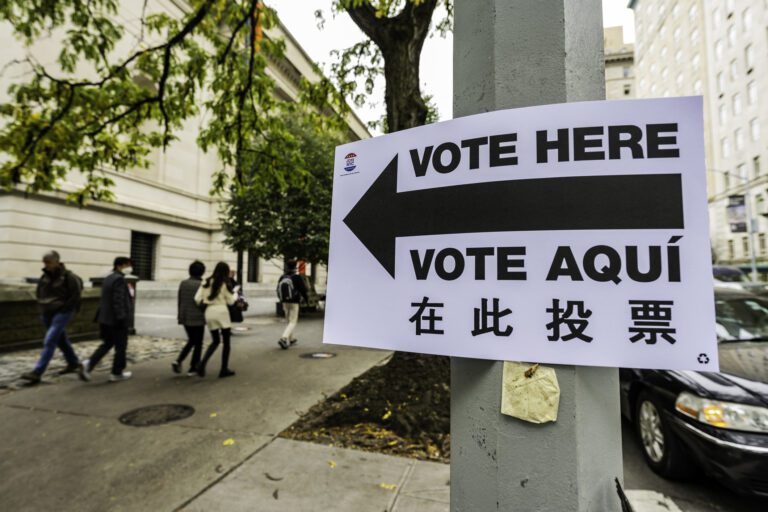 A white sign on a gray post on the side of the street with the words "Vote Here" and "Vote Aqui" with a black arrow pointing to the left